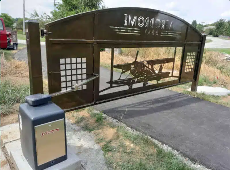 Curved metal gate with an airplane silhouette cutout, opening onto a paved path; an automated barrier box sits to the left.