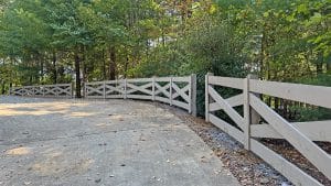 Curved paved path lined with a light gray wooden fence with cross-braces, set in a wooded area with dense trees behind it.